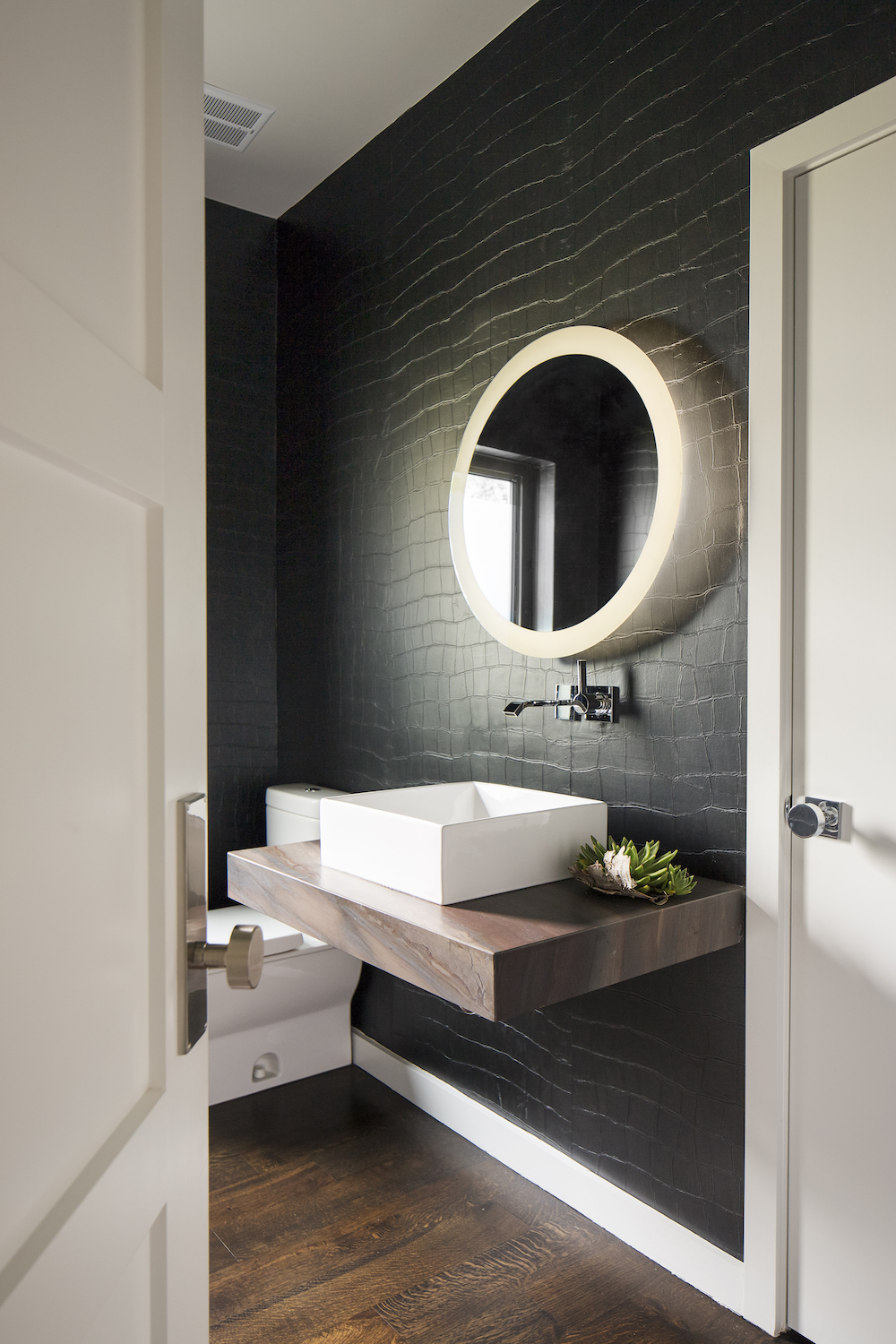 Contemporary powder bathroom interior design with circular mirror, crocodile inspired dark wallcovering and modern fixtures.