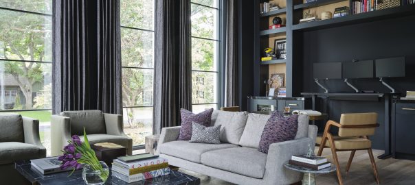 A dramatic home office designed by Pulp Design Studios features floor-to-ceiling steel-framed windows, dark charcoal walls, built-in bookshelves styled with personal objects, a working desk with dual monitors, and a sculptural gold pendant light. A marble coffee table anchors a seating area with a neutral linen sofa and olive velvet chairs, layered over a cowhide rug on light oak floors.