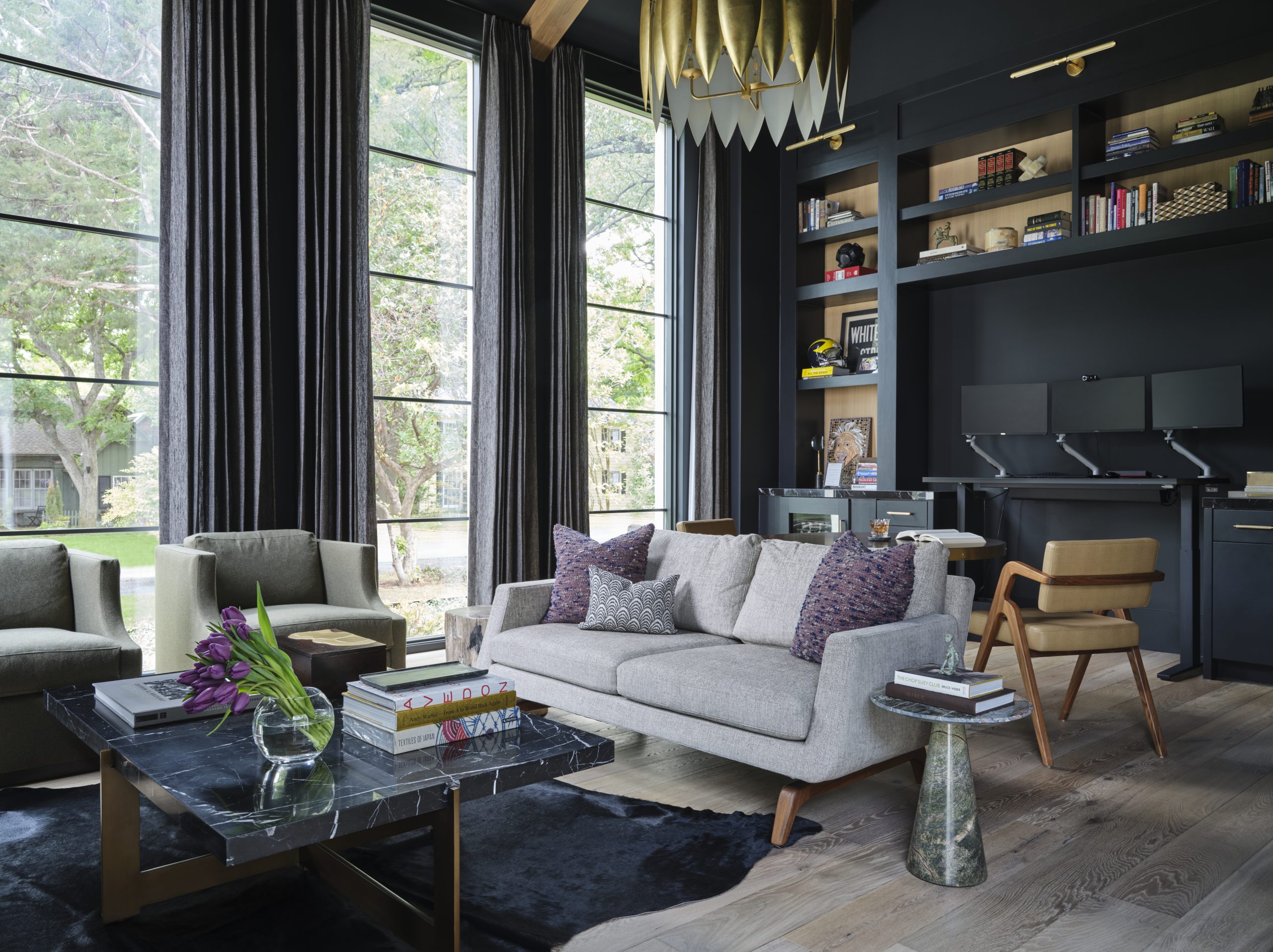 A dramatic home office designed by Pulp Design Studios features floor-to-ceiling steel-framed windows, dark charcoal walls, built-in bookshelves styled with personal objects, a working desk with dual monitors, and a sculptural gold pendant light. A marble coffee table anchors a seating area with a neutral linen sofa and olive velvet chairs, layered over a cowhide rug on light oak floors.